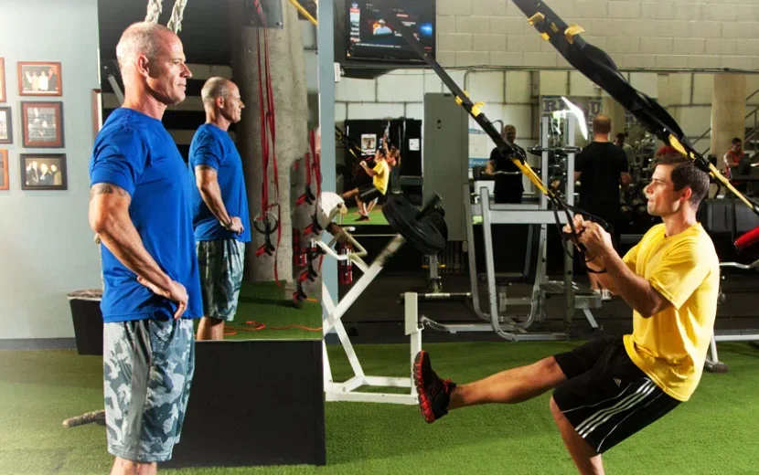 Craig Chapin on the training floor in a blue shirt, hands behind his back, watching a client lean back into a TRX row beside him