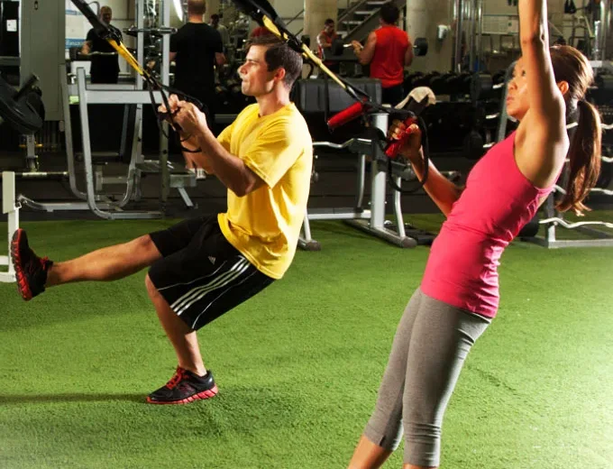 Two clients mid-set on the training turf: one leaning back into a TRX row, the other pressing overhead